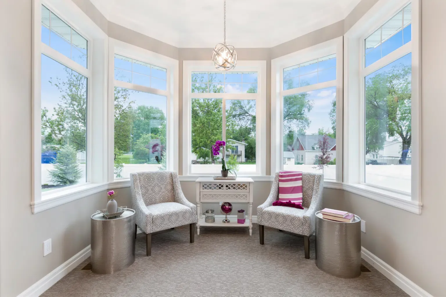 A bay window in a well lit home. The walls are grey and the carpet and furniture match.
